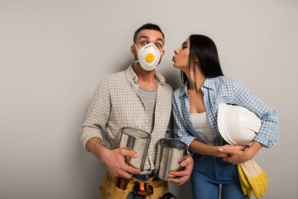 casal de trabalhadores manuais felizes em máscara de segurança segurando latas de tinta e capacete e beijando no cinza
  - Foto, Imagem