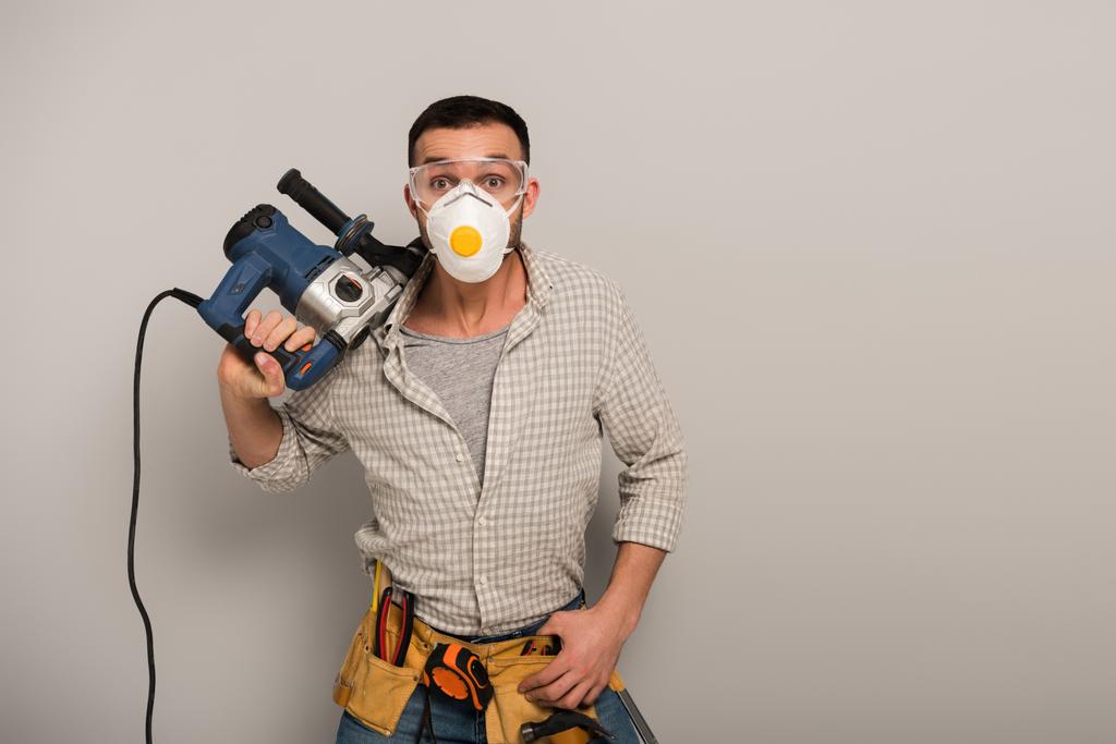 trabajador manual emocional en máscara de seguridad con correa de herramientas que sostiene el taladro eléctrico en gris
 - Foto, Imagen