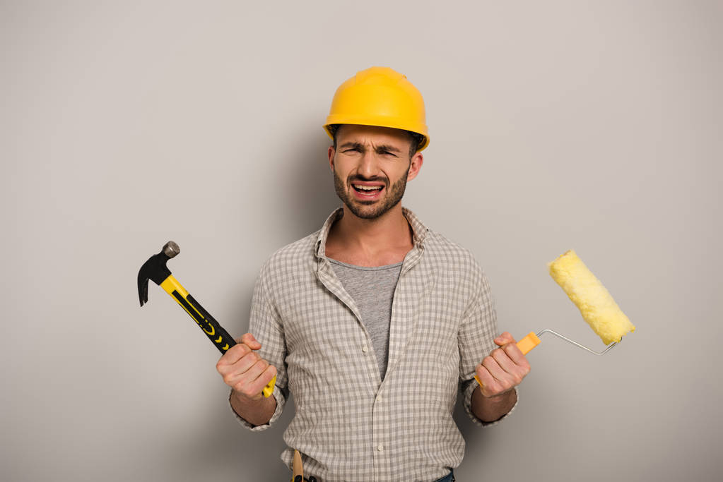 trabajador manual estresado en hardhat sosteniendo rodillo de pintura y martillo en gris
  - Foto, imagen