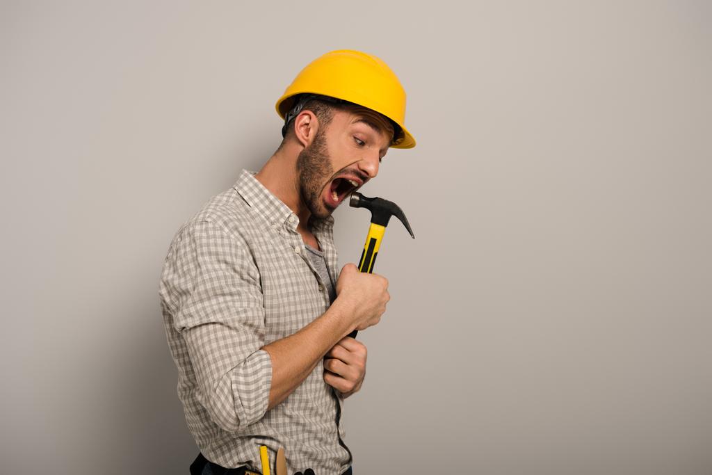 Эмоциональный ремонтник в желтом шлеме кусает серый молоток
   - Фото, изображение