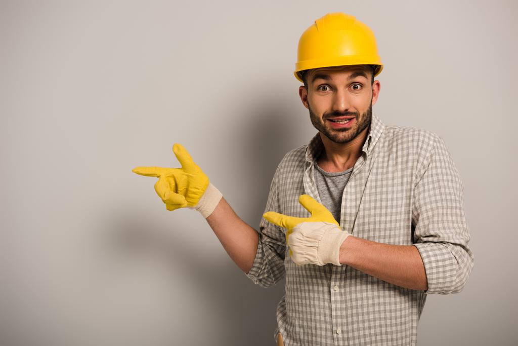 surprised workman in helmet and safety gloves pointing on grey  - Photo, Image