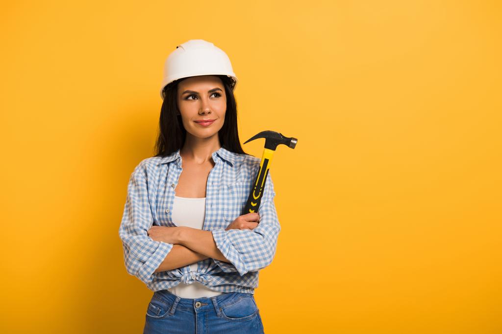 θετική εργάτρια σε κράνος κρατώντας σφυρί με σταυρωμένα χέρια σε κίτρινο  - Φωτογραφία, εικόνα