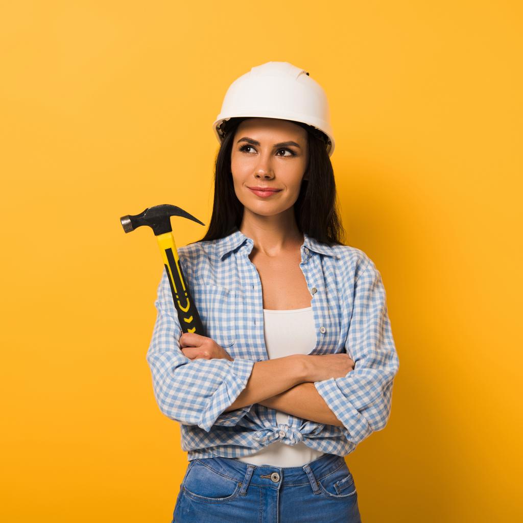 fröhliche Arbeiterin im Helm mit Hammer und verschränkten Armen auf gelb  - Foto, Bild