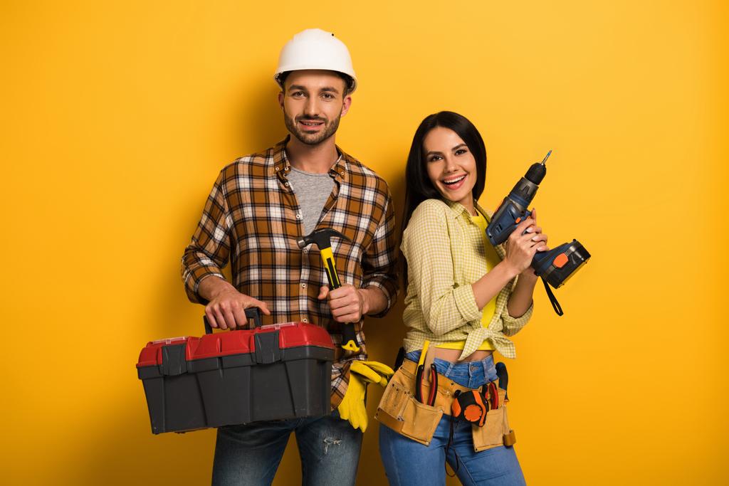 Lächelnde Handarbeiter mit Werkzeugkiste und Elektrobohrer auf gelb    - Foto, Bild