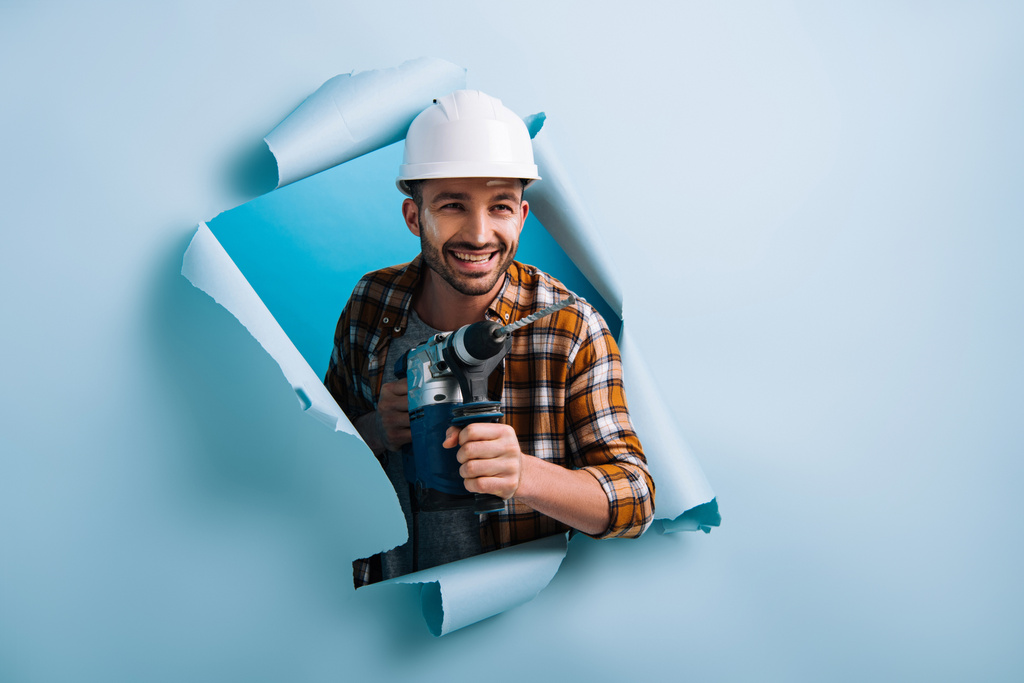 usmívající se údržbář v helmě držící elektrický vrták v roztrhaném papíru, izolovaný na modré - Fotografie, Obrázek
