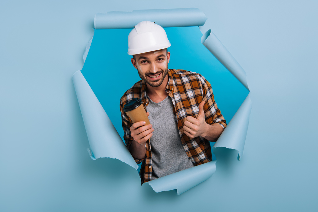 reparador feliz en casco sosteniendo café para ir y mostrando el pulgar hacia arriba en papel roto, aislado en azul
 - Foto, Imagen