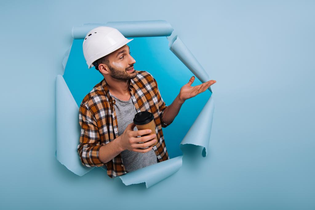 foreman in hardhat holding coffee to go in torn paper, isolated on blue - Photo, Image