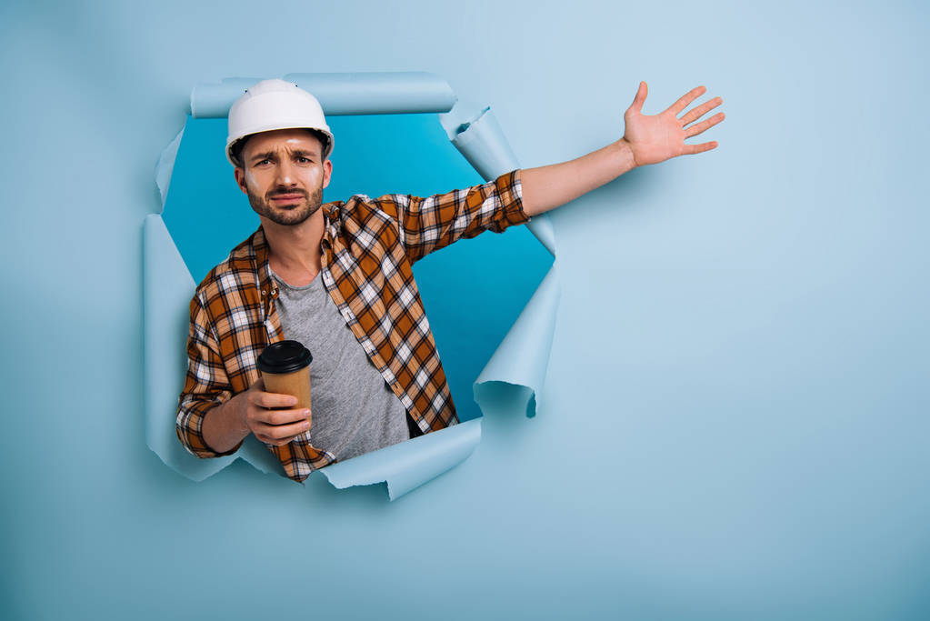 разочарованный мастер в шлеме держит кофе, чтобы пойти в рваной бумаге, изолированные на синий
 - Фото, изображение