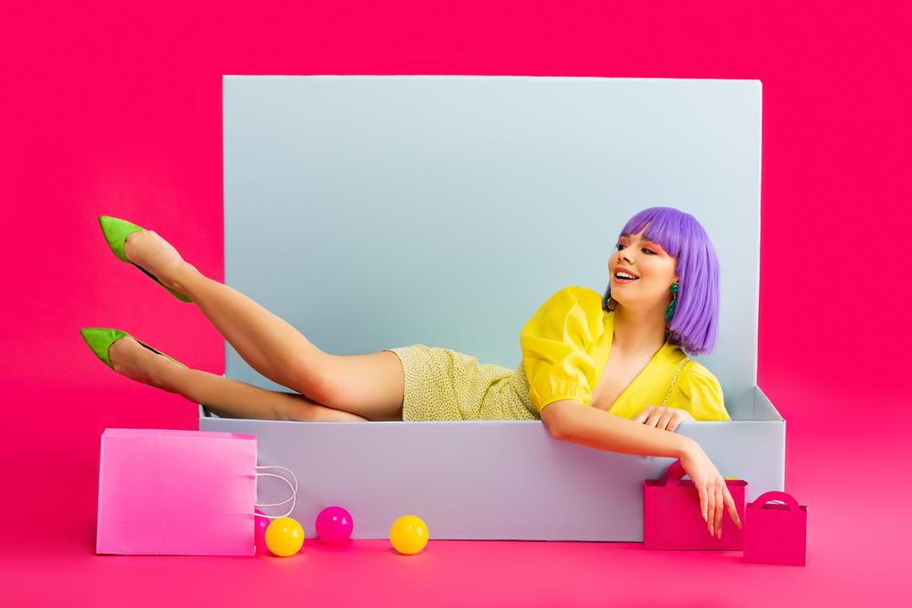 fröhliches Mädchen mit lila Perücke als Puppe in blauer Schachtel mit Einkaufstaschen und Bällen liegend, auf rosa - Foto, Bild