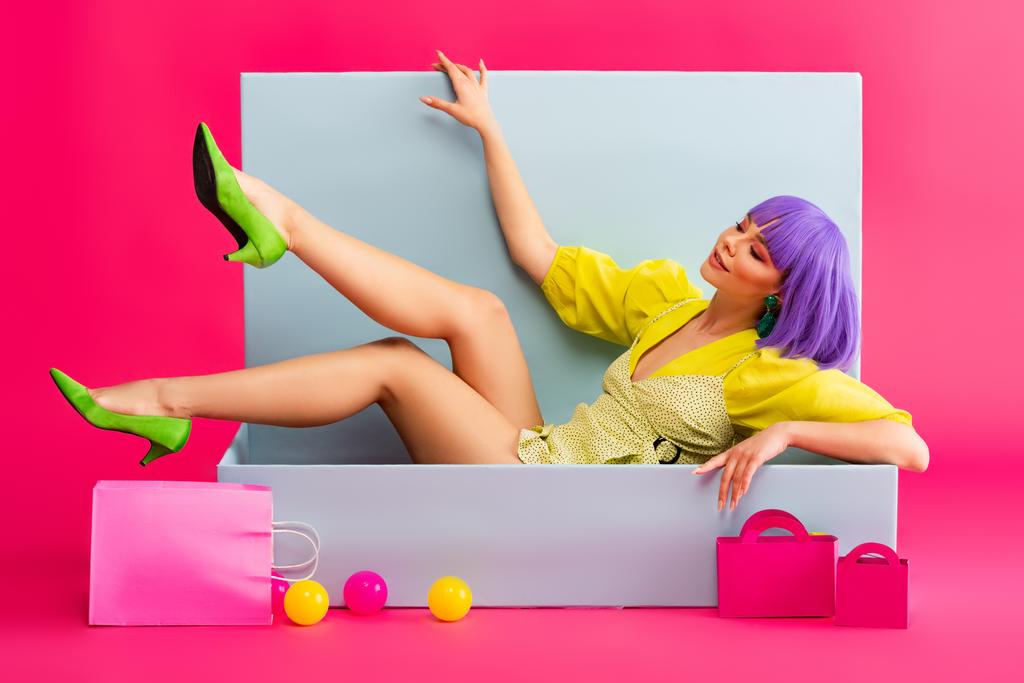 smiling woman in purple wig as doll lying in blue box with shopping bags and balls, on pink - Photo, Image