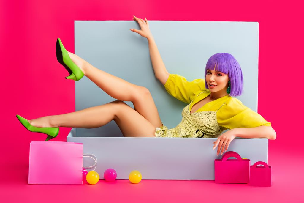 beautiful emotional girl in purple wig as doll lying in blue box with shopping bags and balls, on pink - Photo, Image