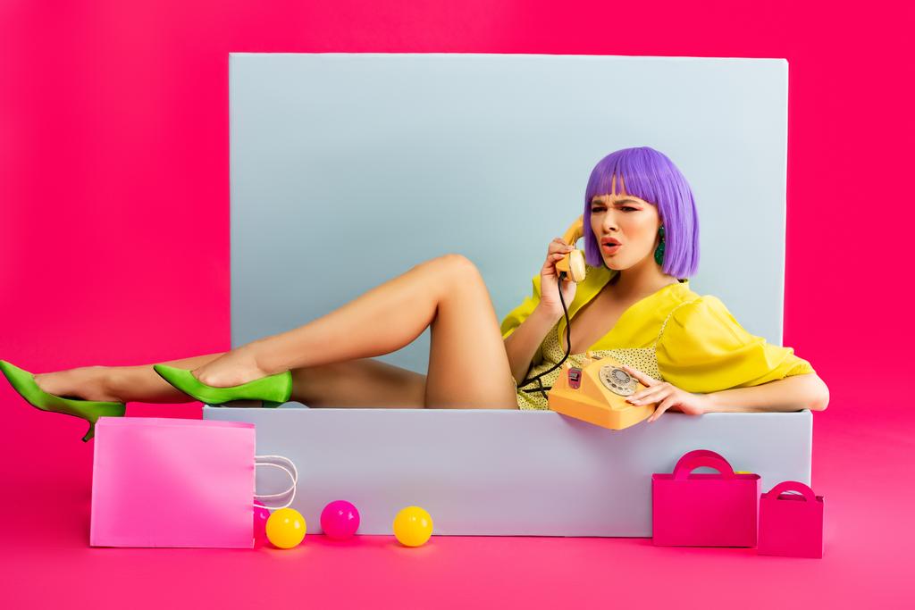aggressive girl in purple wig as doll talking on vintage phone while sitting in blue box with balls and shopping bags, on pink - Photo, Image