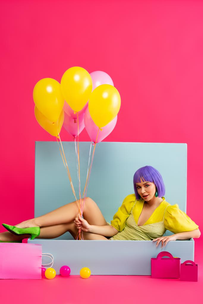 mulher atraente em peruca roxa como boneca segurando balões e sentado em caixa azul com bolas e sacos de compras, em rosa
 - Foto, Imagem