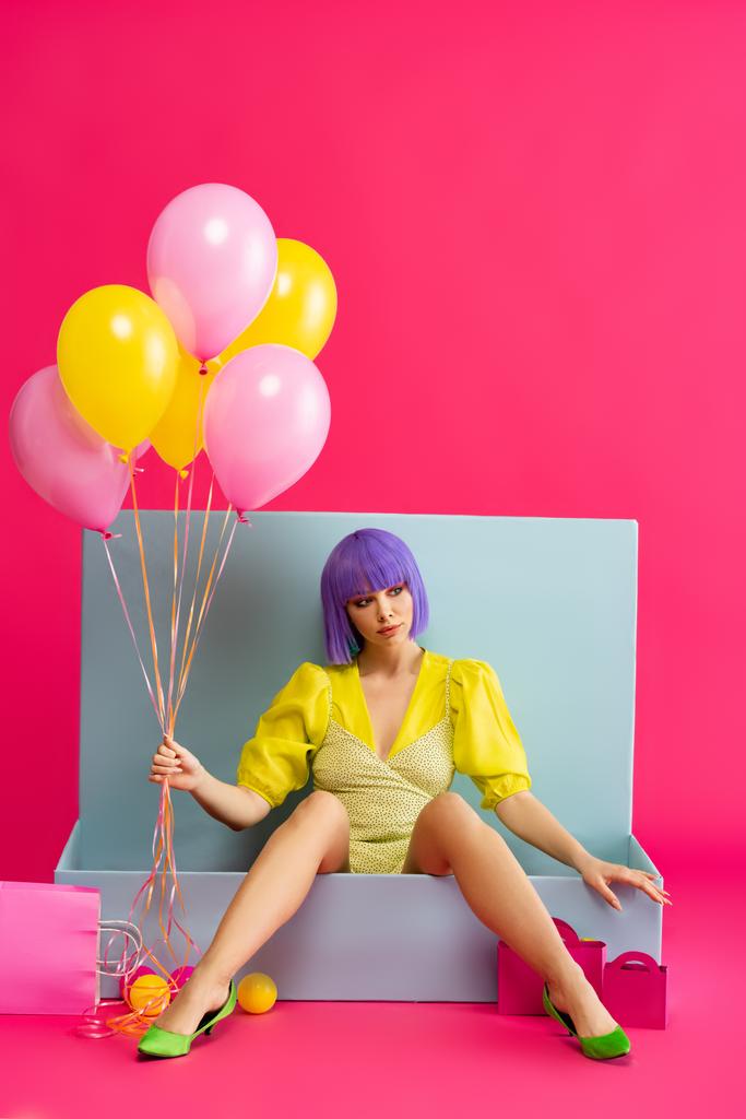 bella ragazza in parrucca viola come bambola che tiene palloncini e seduto in scatola blu con palle e borse della spesa, su rosa
 - Foto, immagini
