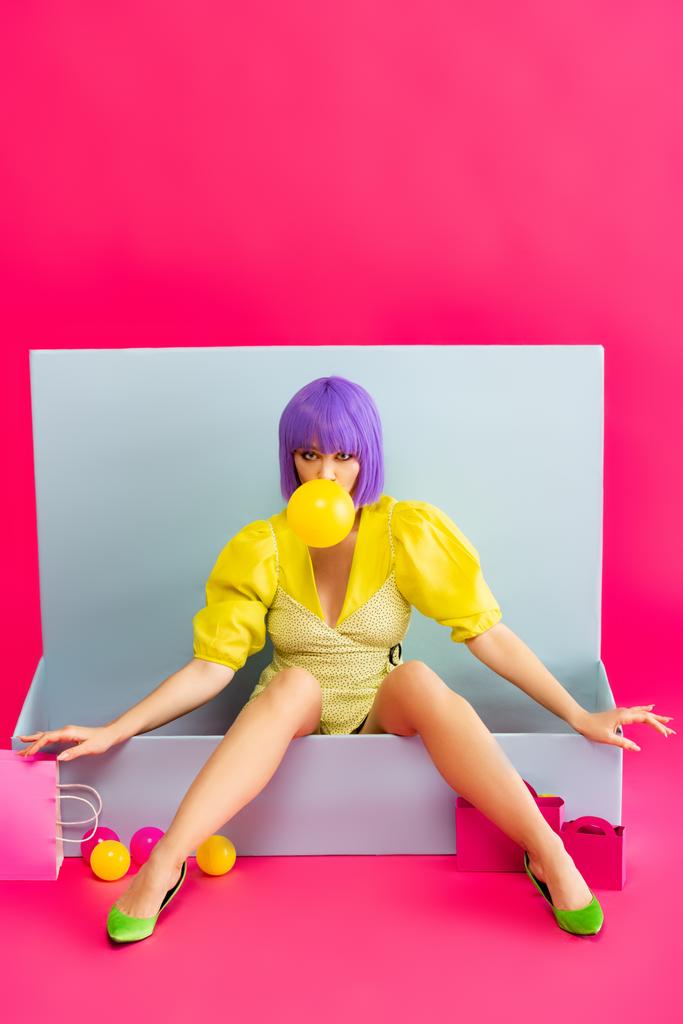 girl in purple wig as doll blowing bubble gum while sitting in blue box with balls and shopping bags, on pink - Photo, Image