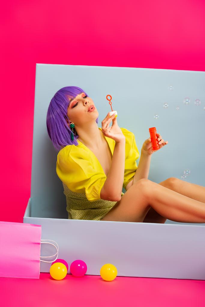 beautiful girl in purple wig as doll blowing soap bubbles while sitting in blue box with balls and shopping bag, on pink - Photo, Image