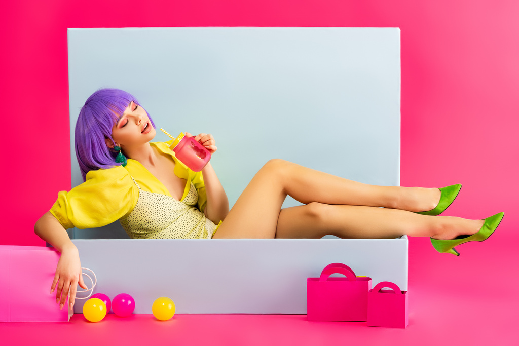 schönes Mädchen in lila Perücke als Puppe, die aus Glas trinkt, während sie in einer blauen Schachtel mit Bällen und Einkaufstaschen sitzt, auf rosa - Foto, Bild