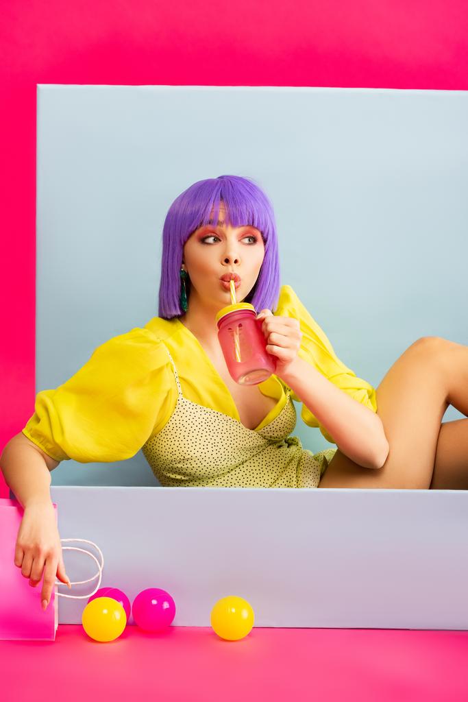 Pop-Art-Mädchen mit lila Perücke als Puppe, die aus Glas trinkt, während sie in blauer Schachtel mit Bällen und Einkaufstasche sitzt, auf rosa - Foto, Bild