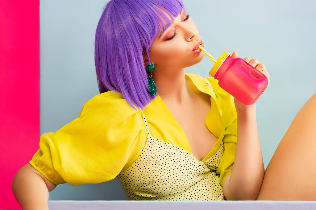 beautiful pop art girl in purple wig as doll drinking from jar while sitting in blue box, isolated on pink - Photo, Image