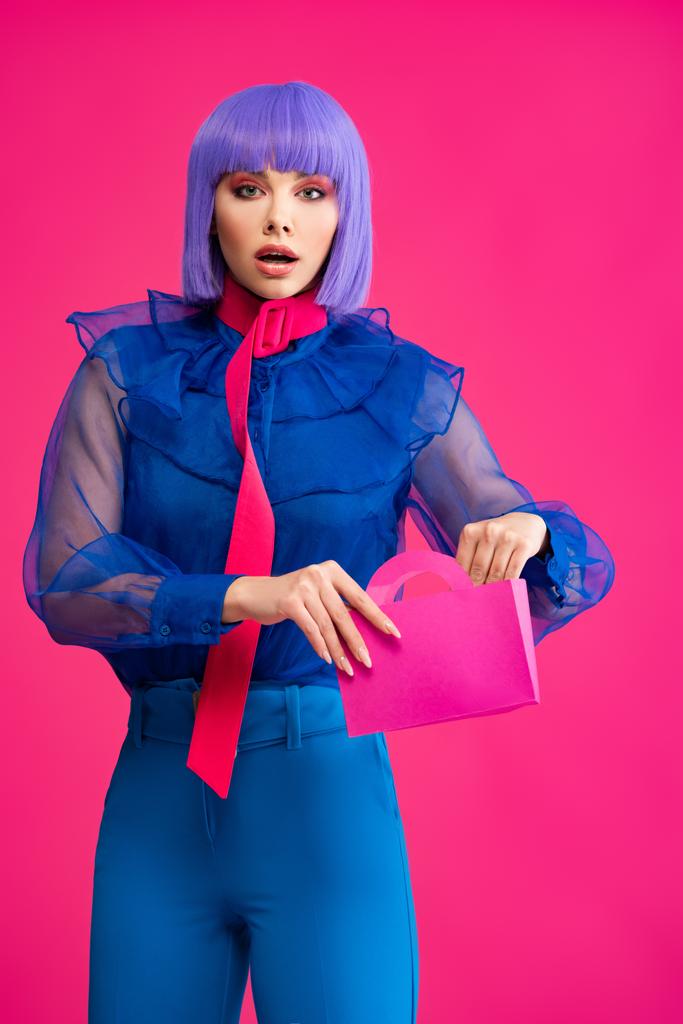 κομψό σοκαρισμένο κορίτσι σε μωβ περούκα κρατώντας μια μικρή τσάντα για ψώνια, απομονωμένο σε ροζ - Φωτογραφία, εικόνα