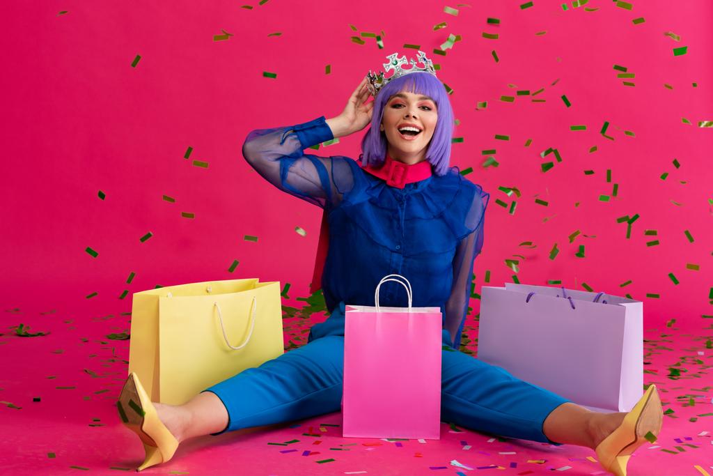 chica feliz en peluca púrpura y corona sentado con bolsas de compras y confeti de vacaciones, en rosa
 - Foto, imagen