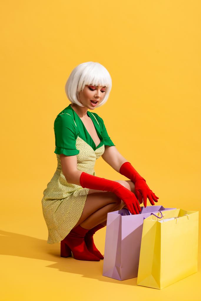 surprised pop art girl in white wig looking into shopping bags, on yellow - Photo, Image