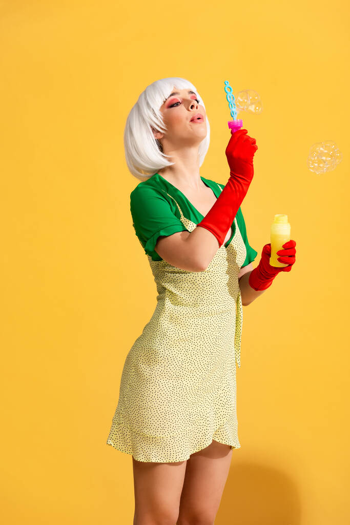 mooi pop art meisje in wit pruik blazen zeepbellen, op geel  - Foto, afbeelding