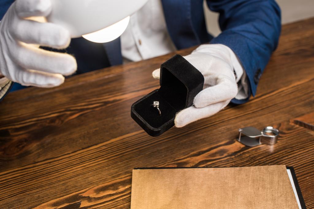 Обрезанный вид ювелира оценщик держа кольцо с драгоценным камнем в коробке возле лампы за столом изолированы на сером
 - Фото, изображение