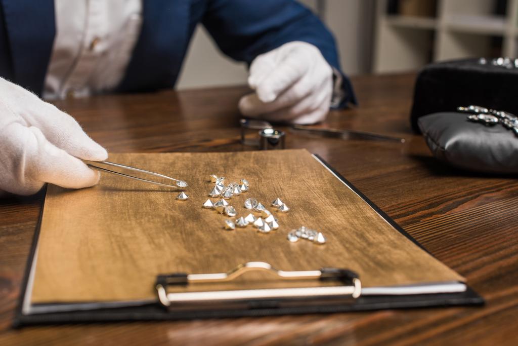 Cropped view of jewelry appraiser with pliers examining gemstones on wooden board on table in workshop - Photo, Image