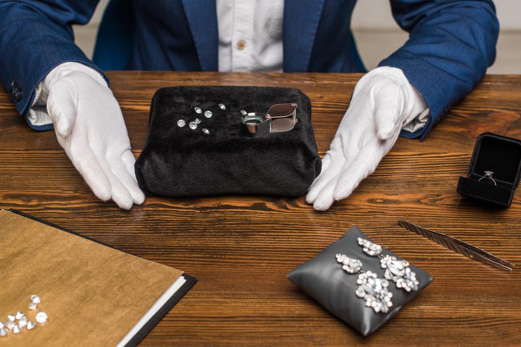 Vue recadrée de l'évaluateur de bijoux dans des gants près de pierres précieuses près de bijoux sur une table en bois
  - Photo, image