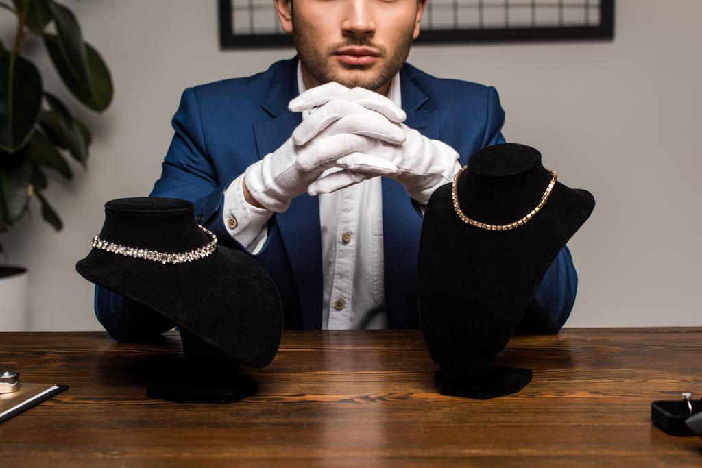 Gesneden weergave van sieraden taxateur in de buurt van kettingen op sokkels op tafel in de werkplaats - Foto, afbeelding