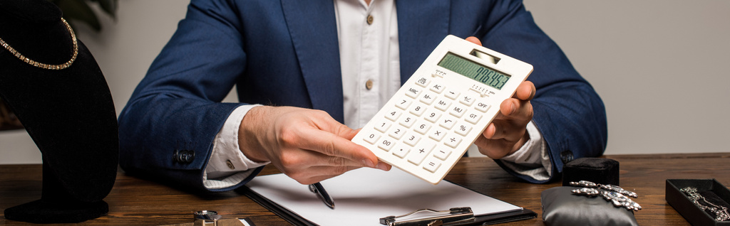 Обрезанный вид ювелирного оценщика с калькулятором возле буфера обмена и ювелирных изделий на столе, панорамный снимок
 - Фото, изображение