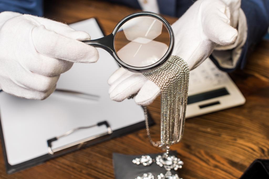 Vista ritagliata di gioielliere in possesso di collana e lente d'ingrandimento vicino alla calcolatrice, appunti e orecchini sul tavolo
 - Foto, immagini