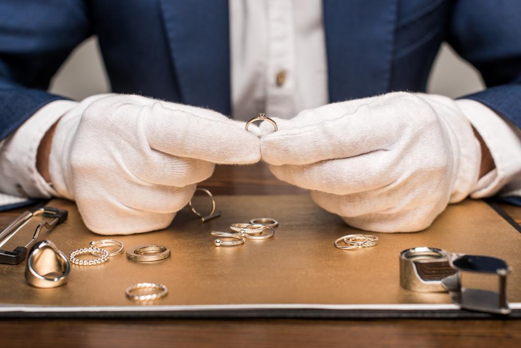 Vista recortada del tasador de joyas que sostiene el anillo de joyería cerca de la junta y lupa en la mesa aislada en gris
 - Foto, Imagen