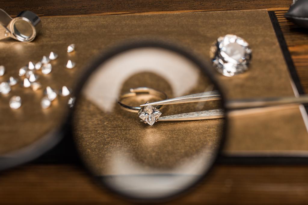 Concentration sélective de loupe, bague de bijoux avec pierre précieuse dans des pinces à épiler à bord sur une table en bois
 - Photo, image