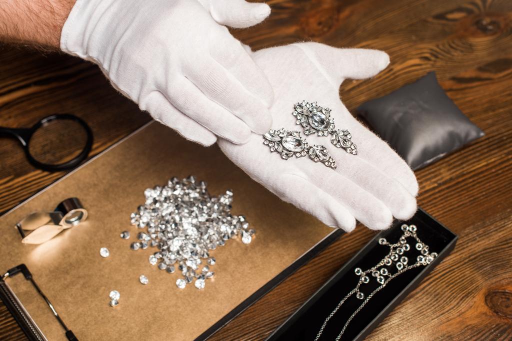 Vista recortada del tasador de joyas que sostiene pendientes cerca de piedras preciosas y lupas a bordo en la mesa
 - Foto, imagen