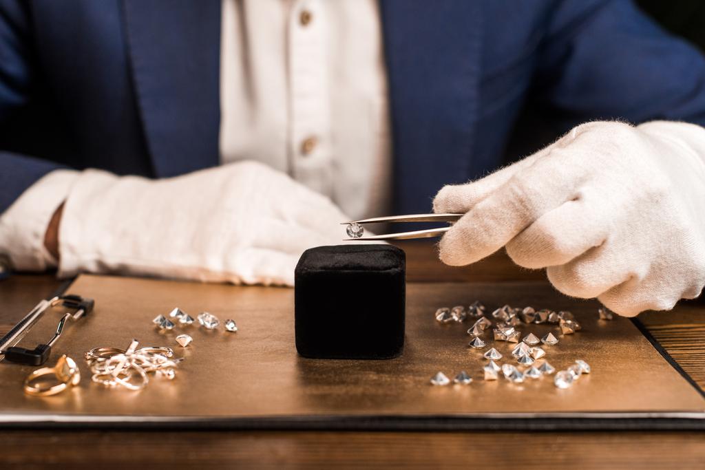 Oříznutý pohled na klenotnictví odhadce držení drahokam v pinzetě v blízkosti šperky na palubě na stole izolované na černé  - Fotografie, Obrázek