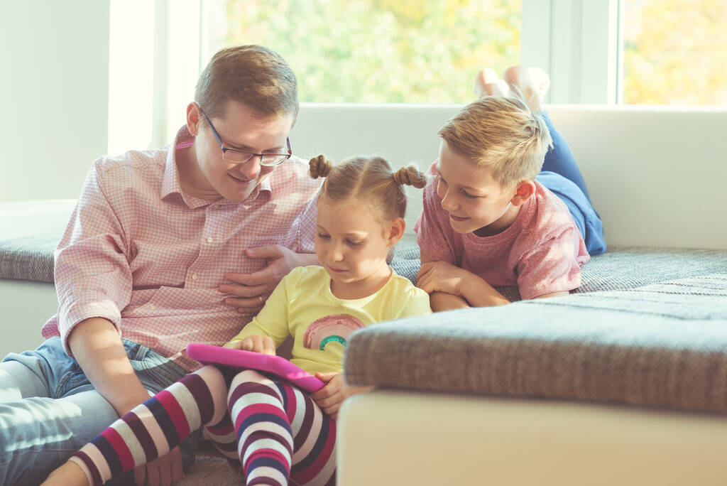 Щасливий молодий красивий батько розважається зі своїми двома милими дітьми, граючи радісно з планшетом
 - Фото, зображення