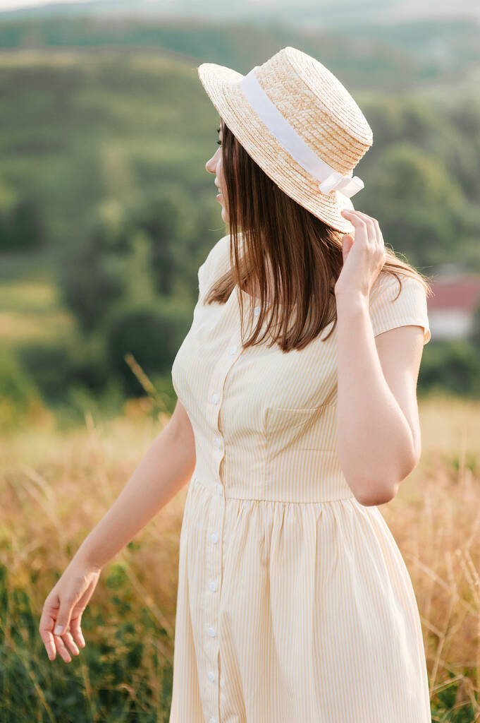 Brunette meisje in een gele jurk en een hoed op de natuur. Wandelingen op een veld op een heuvel. - Foto, afbeelding