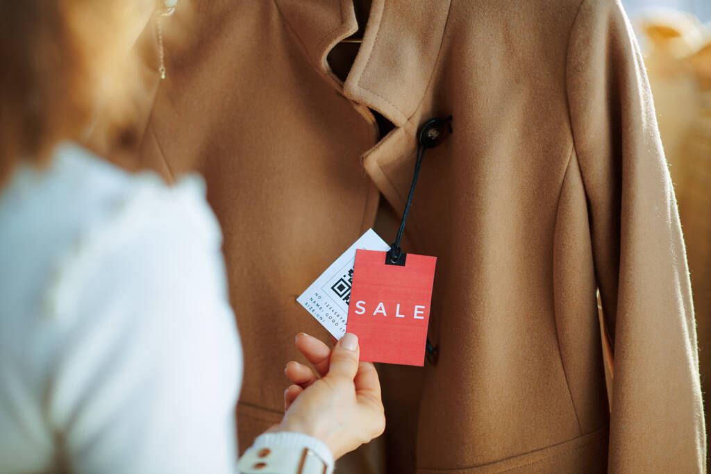 Close-up op trendy vrouw in witte trui en rok in moderne modewinkel onderzoekt verkoop prijskaartje op beige jas. - Foto, afbeelding