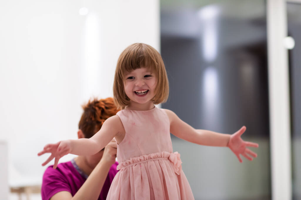 家庭で楽しい夜を過ごしてる若いお母さんかわいい娘を助けながらピンクのドレスを着て家で踊る - 写真・画像