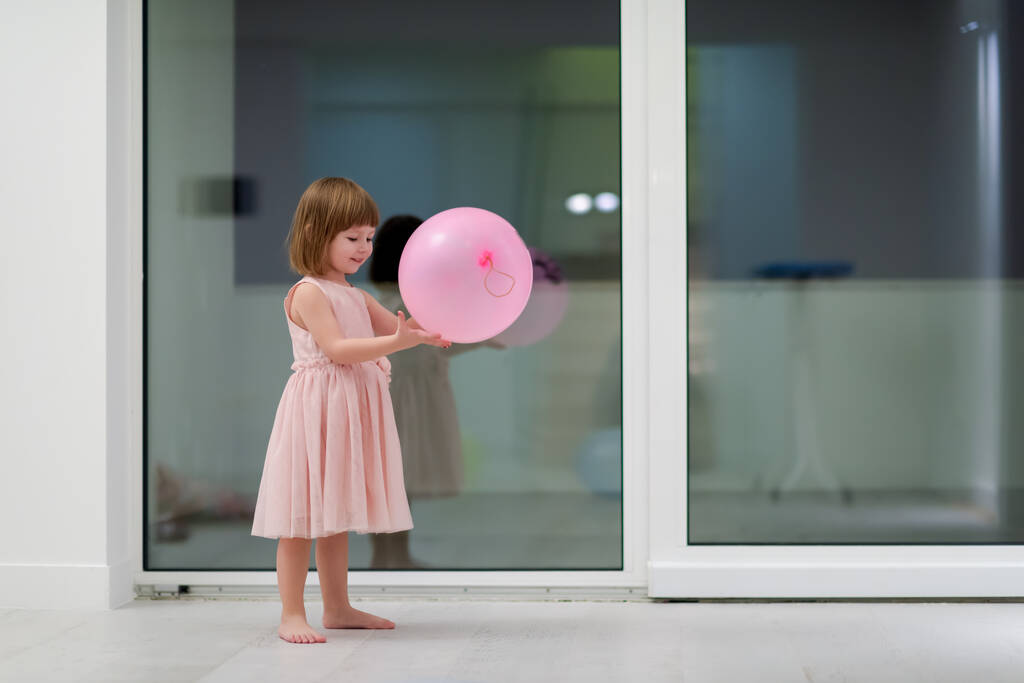 šťastný roztomilý holčička v růžové šaty těší při hraní s balónky na krásný večer v blízkosti okna doma - Fotografie, Obrázek