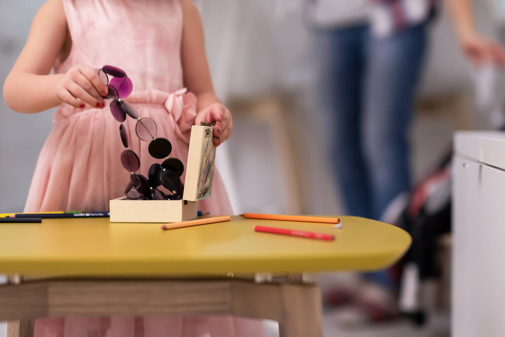 feliz familia pasando tiempo juntos en casa linda hijita en un vestido rosa jugando y pintando el joyero mientras joven pelirroja madre planchando ropa detrás de ella
 - Foto, Imagen