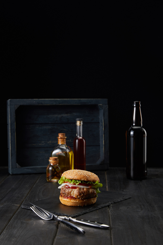 Burger auf dunkler Holzoberfläche, Gabel, Messer und Ölflaschen isoliert auf schwarz - Foto, Bild