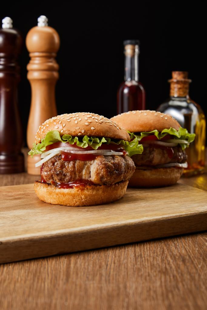 selective focus of two burgers on wooden cutting board with oil, vinegar and beer bottles, pepper and salt mills isolated on black - Photo, Image