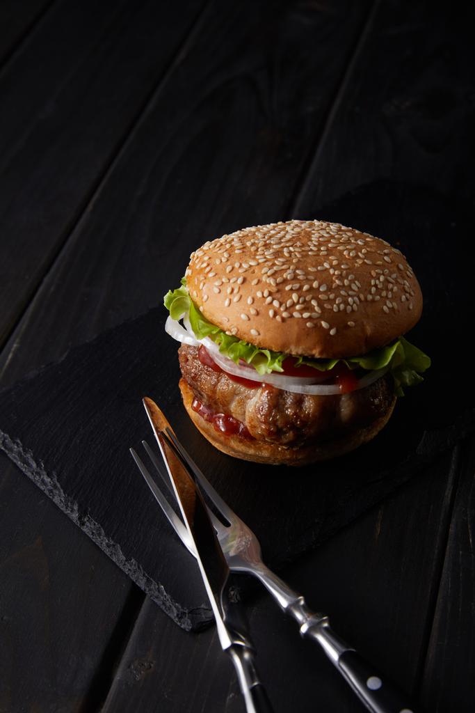 high angle view of a burger, fork and knife on a black wooden surface - Photo, Image