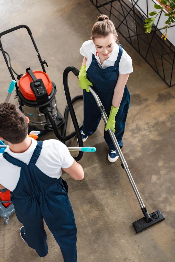 vysoký úhel pohledu na atraktivní čistič vysávání podlahy v blízkosti kolegy vozík s čisticími prostředky - Fotografie, Obrázek