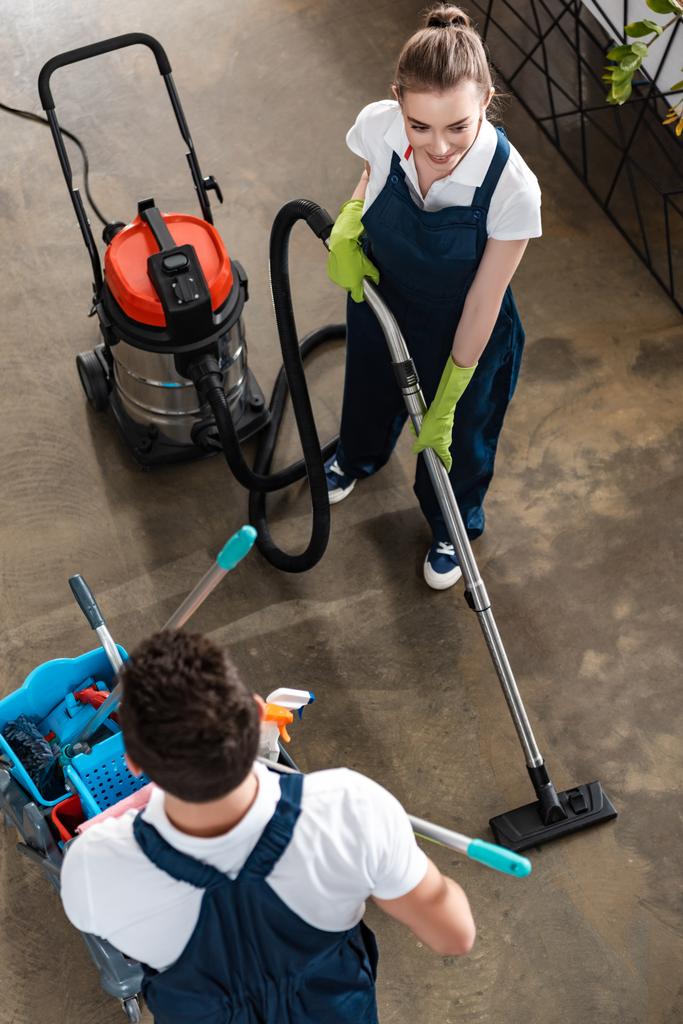 vysoký úhel pohledu na usmívající se čistič vysávání podlahy v blízkosti kolegy nesoucí vozík s čistícími prostředky - Fotografie, Obrázek