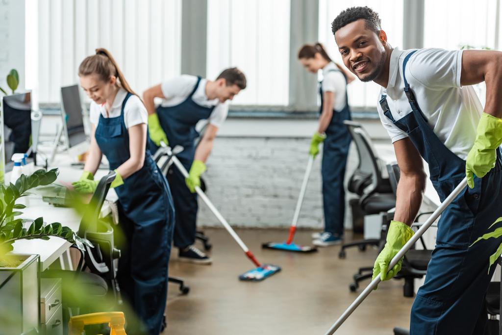 foco seletivo de sorrindo equipe multicultural de limpadores lavando chão com esfregonas no escritório
 - Foto, Imagem