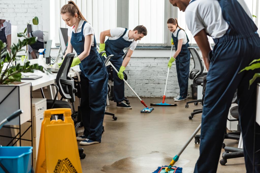 fiatal takarítókból álló multikulturális csapat mossa fel a padlót felmosóval az irodában - Fotó, kép